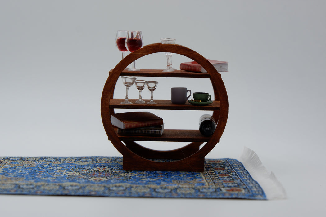 A miniature circular wooden shelf with three tiers, styled with tiny books, wine glasses, a teacup, and a miniature rug on a white studio background.