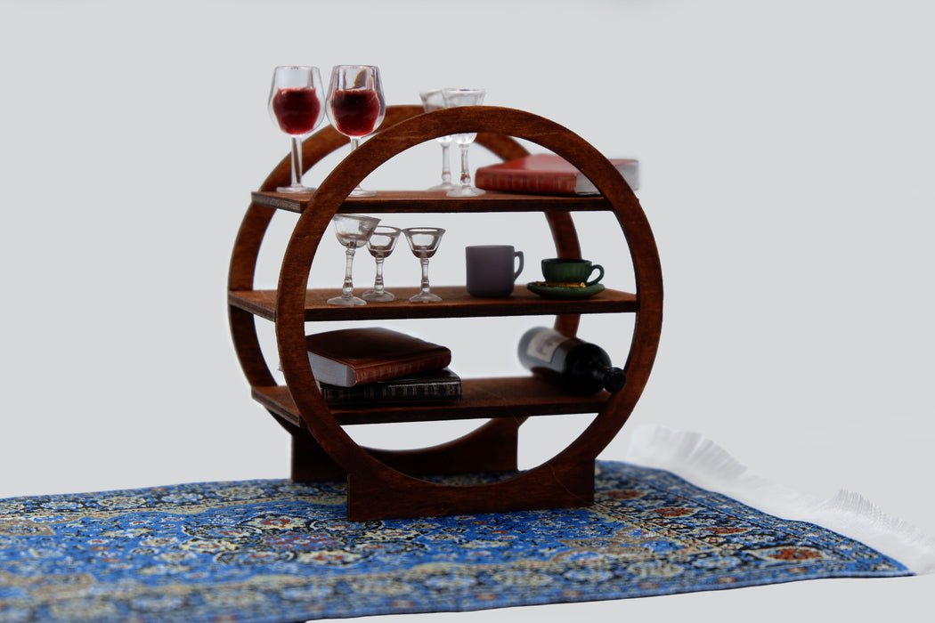 A three-tier wooden serving shelf with a circular frame design, styled with wine glasses, books, and a mug, set on a blue patterned rug against a white background.